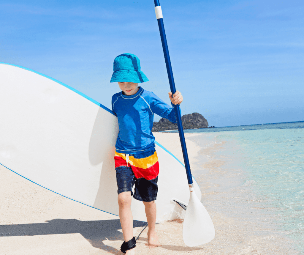 A small boy holds a paddle and paddle board as he walks along a beach.