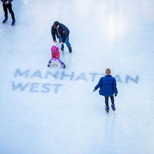 A dad pushes his young daughter on a skating aid, while another skater passes by at the The Rink at Manhattan West.