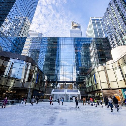 A view of several ice skaters at The Rink at Manhattan West, with surrounding buildings towering overhead.