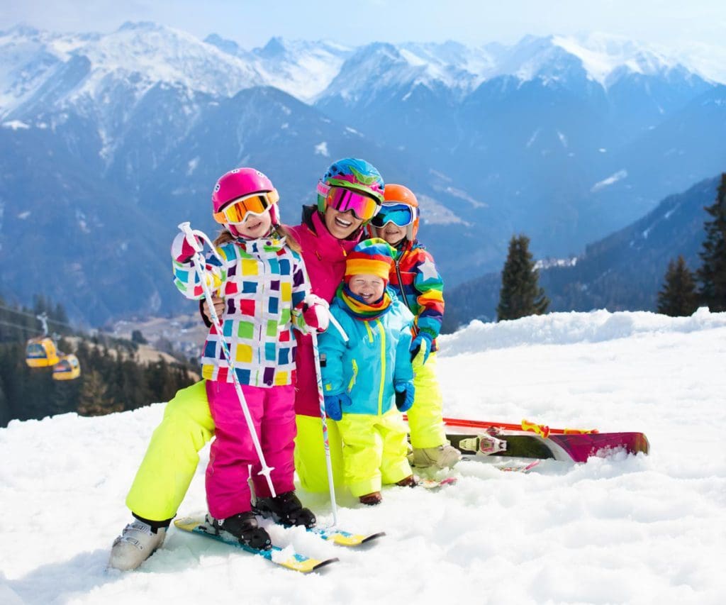 A mom and three kids, all in colorful snow gear, helmets, and skis, enjoy a sunny day on-mountain, searching for kids ski-free programs can help plan a family ski trip on a budget.