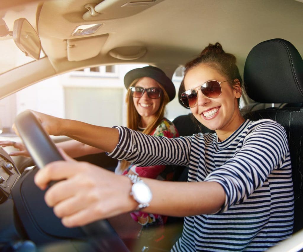 Two woman sit in the front seat of a car, while on a girls' trip.