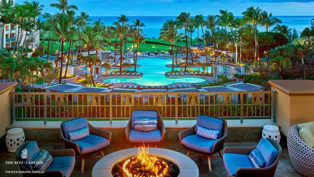 Several empty chairs surround an outdoor bon fire, with the pool at the The Ritz-Carlton Maui, Kapalua in the distance.