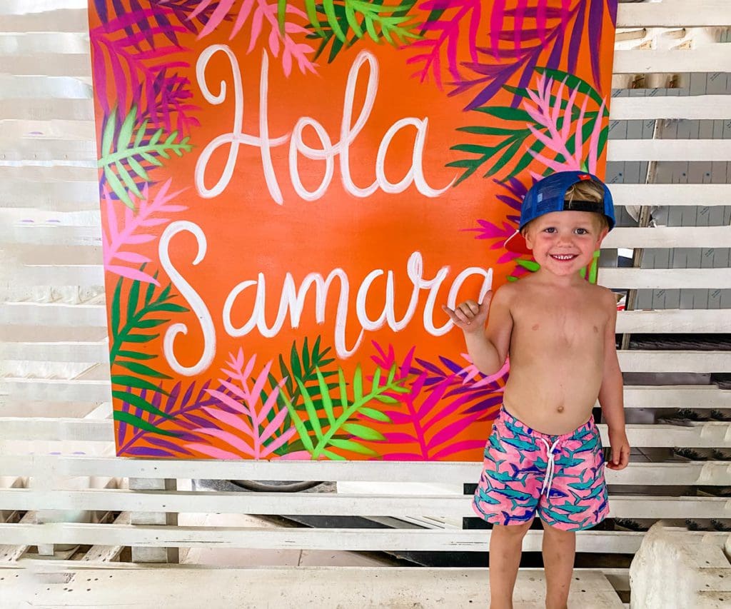 A young boy wearing a swim suit and hat stands next to a vibrant sign reading "Hola Samara".