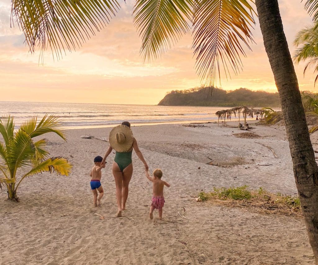 A mom holds the hands of two children, one on each side, as they walk along the beach toward the ocean at sunset.