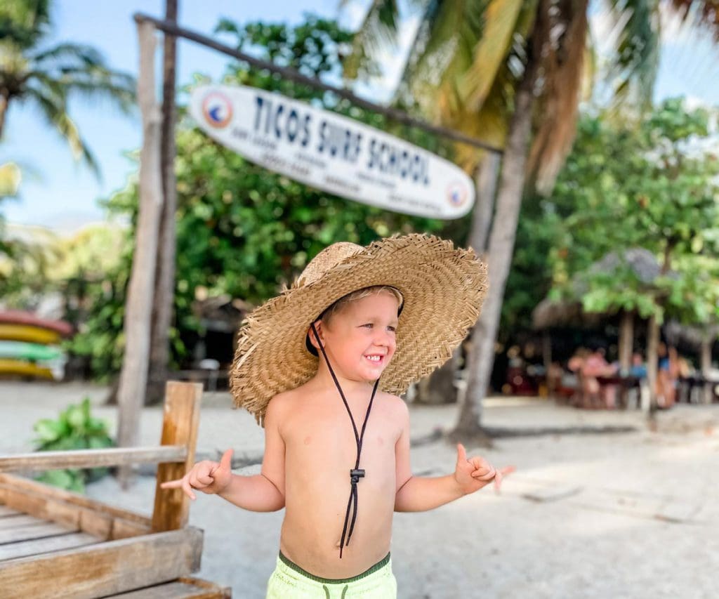A young boy signing 'hang ten' stands on the beach wearing a swim suit and straw hat.
