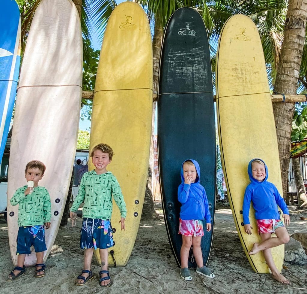 Four young boys stand in front of their own surfboards on the beach.