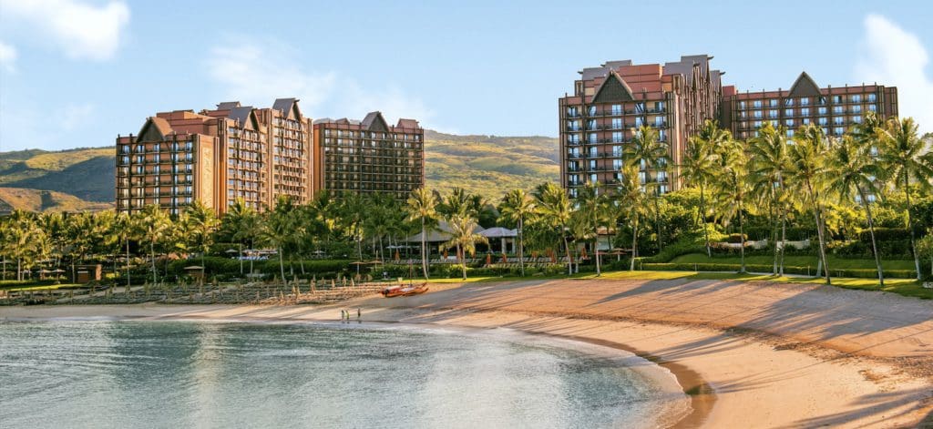 A pristine beach sprawls along the ocean shore, with resort buildings from Aulani, A Disney Resort & Spa in the distance.