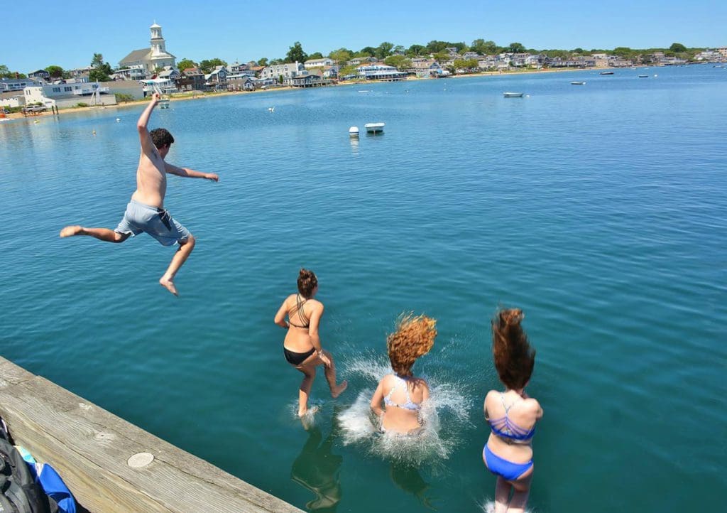 Several kids jump into the water from a break wall in Provincetown on a sunny day.