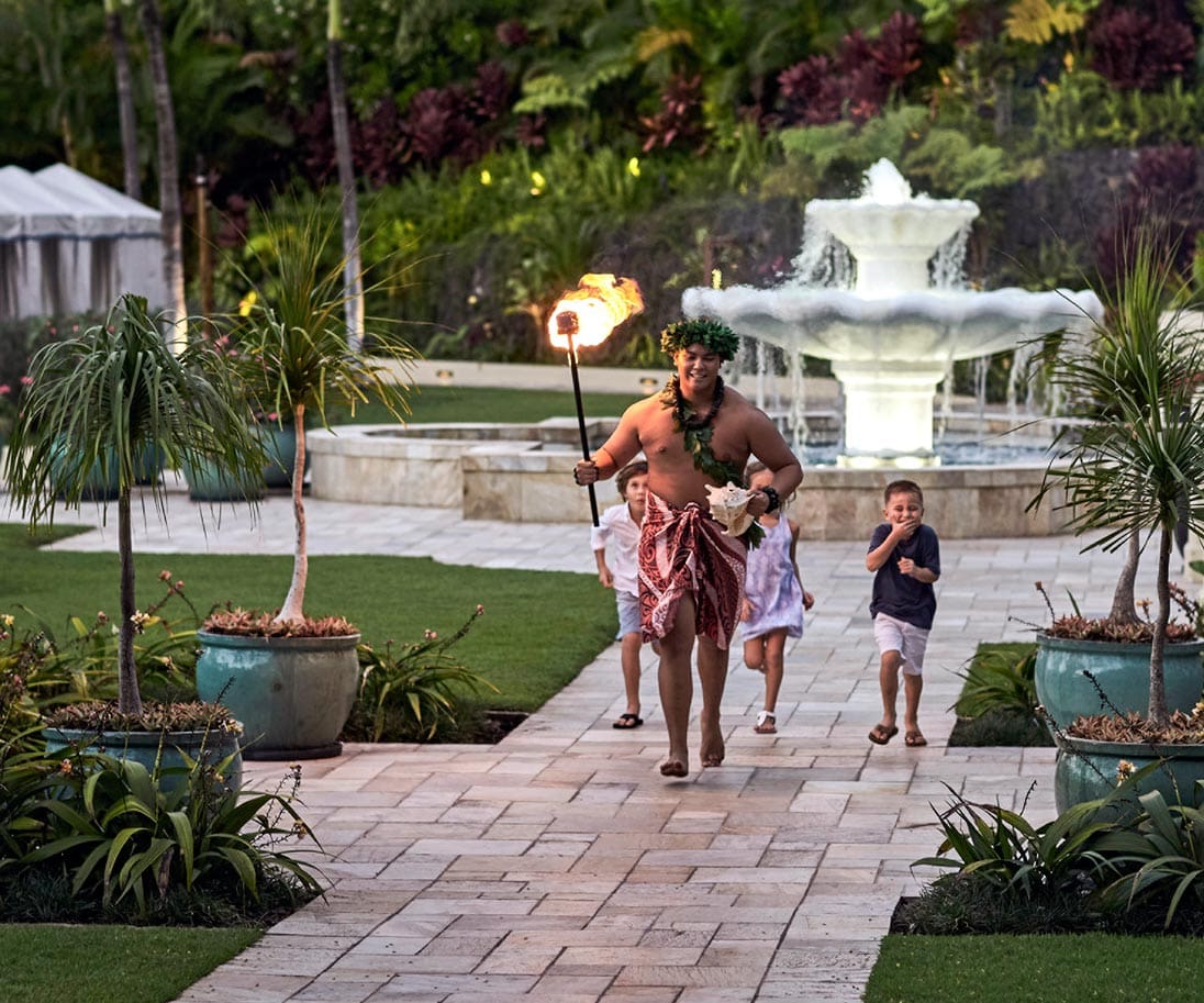 A man in traditional Hawaiin clothes holds a torch and leads some children through the grounds of Four Seasons Resort Maui at Wailea.