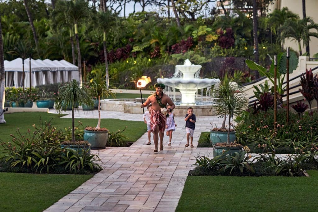 A man in traditional Hawaiian clothes holds a torch and leads some children through the grounds of Four Seasons Resort Maui at Wailea, one of the best luxury hotels in the U.S for families. 