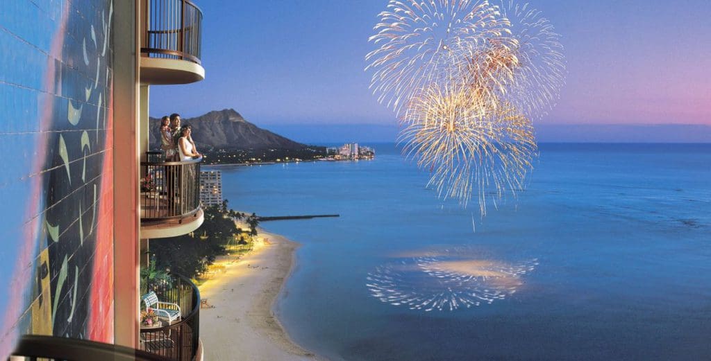 A family stands on a balcony off one of the rooms at Hilton Hawaiian Village Waikiki Beach Resort, enjoying fireworks over the ocean at one of the best resorts for families in O'ahu,