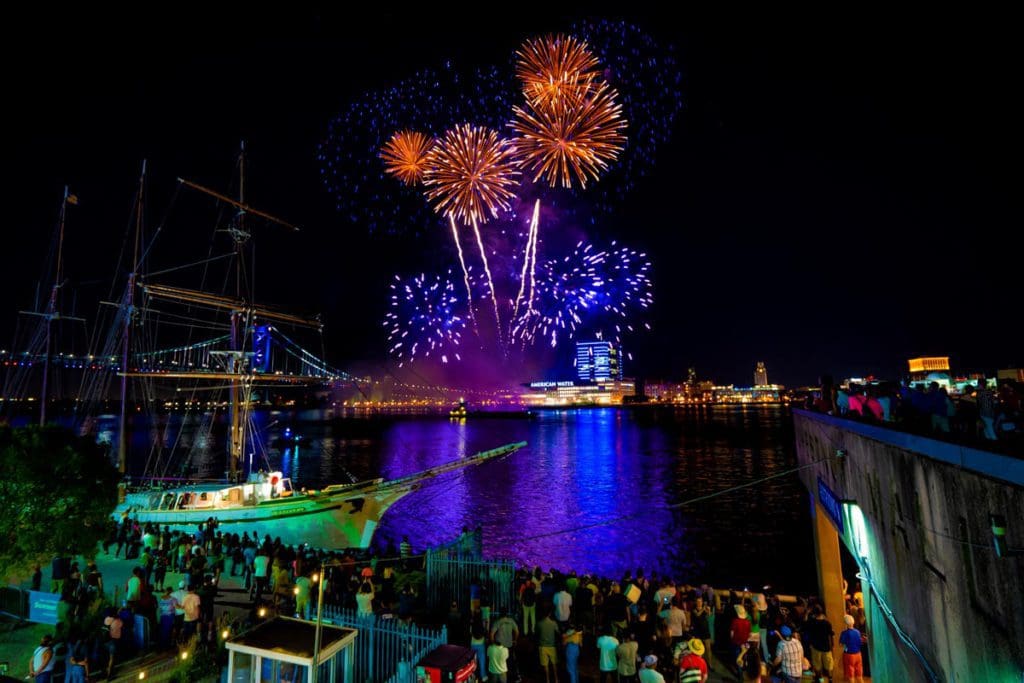 A crowd of people gathers along the river in Philadelphia, enjoying a huge, colorful firework display, one of the best Fourth of July destinations for a family trip.