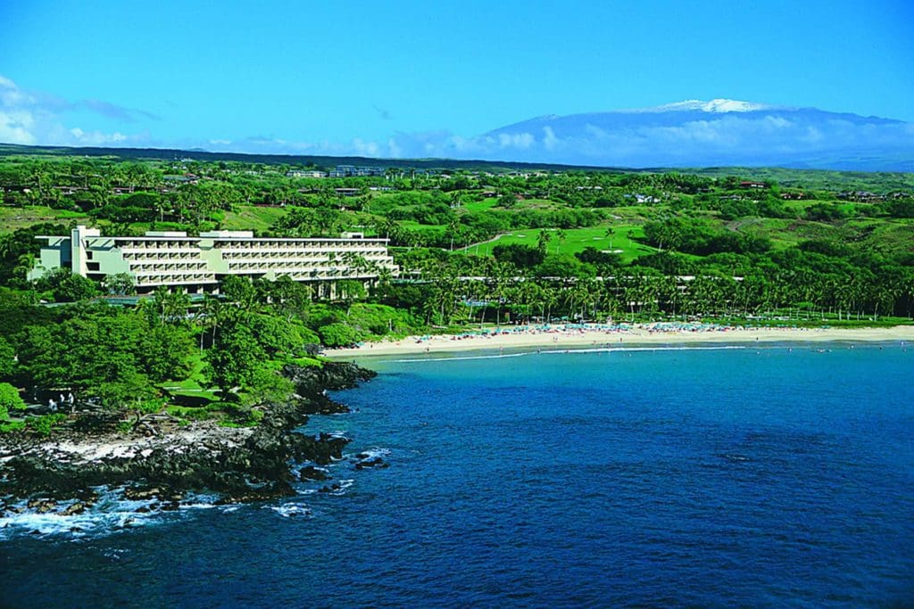 The resort buildings of the Mauna Kea Beach Hotel, Autograph Collection sit above the beach shore and ocean, surrounded by lush greenery.