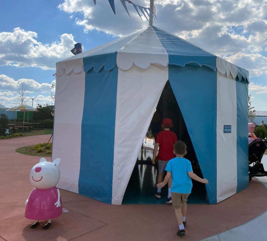 Two kids walk into a blue and white striped tent on a sunny day.