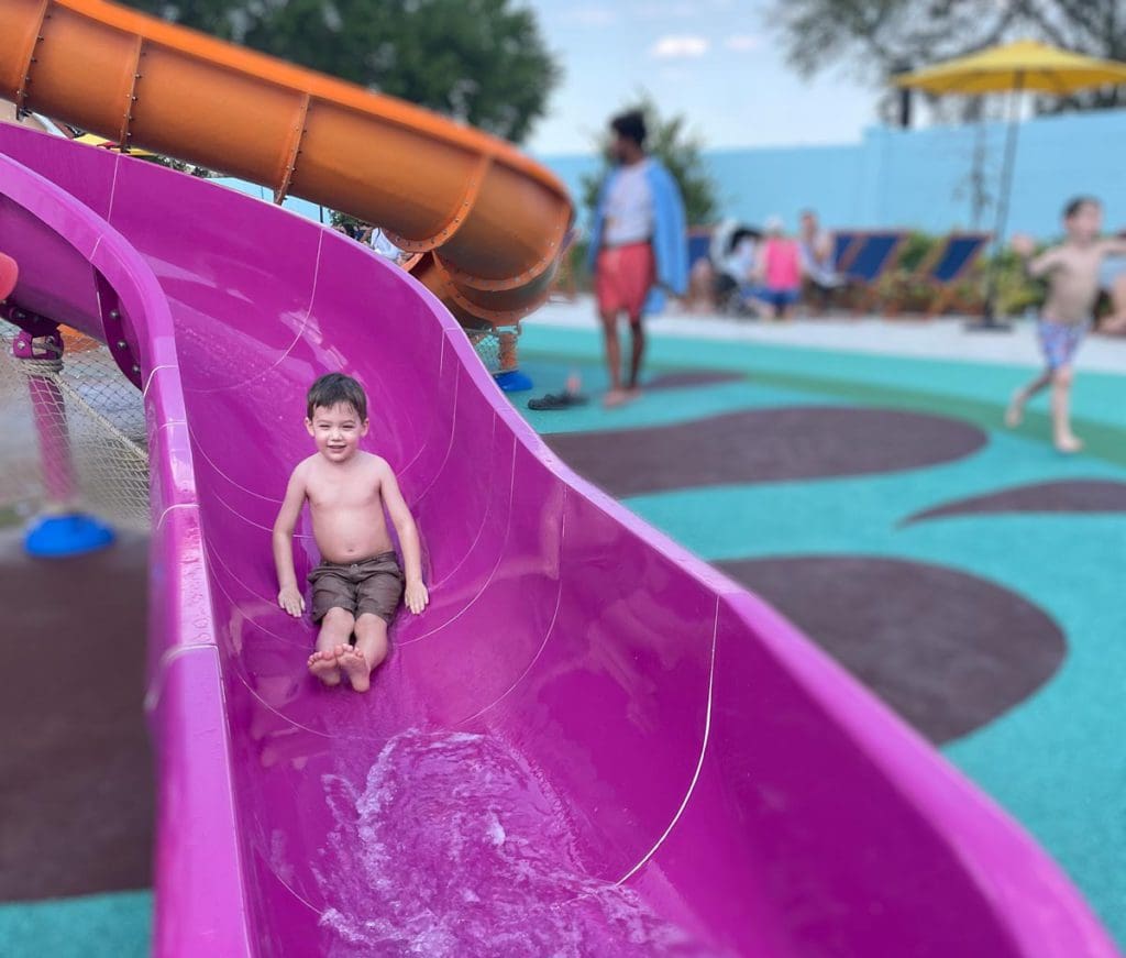 A young boy races down a pink slide at an outdoor splash pad.