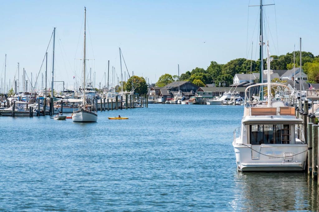 A view of the seaport in Mystic, featuring several docked sail boats, and the shoreline in the distance.