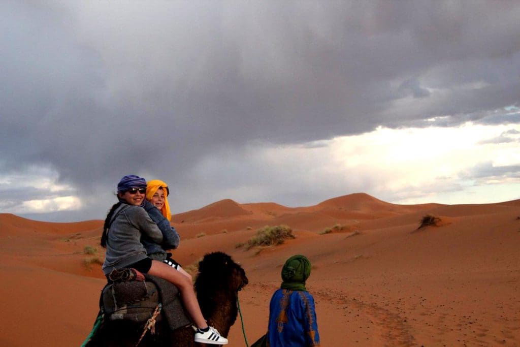 A guide leads a camel, atop of which are a mom and child, in the desert of Morocco, one of the best things to do in our Perfect Morocco Itinerary for Families.