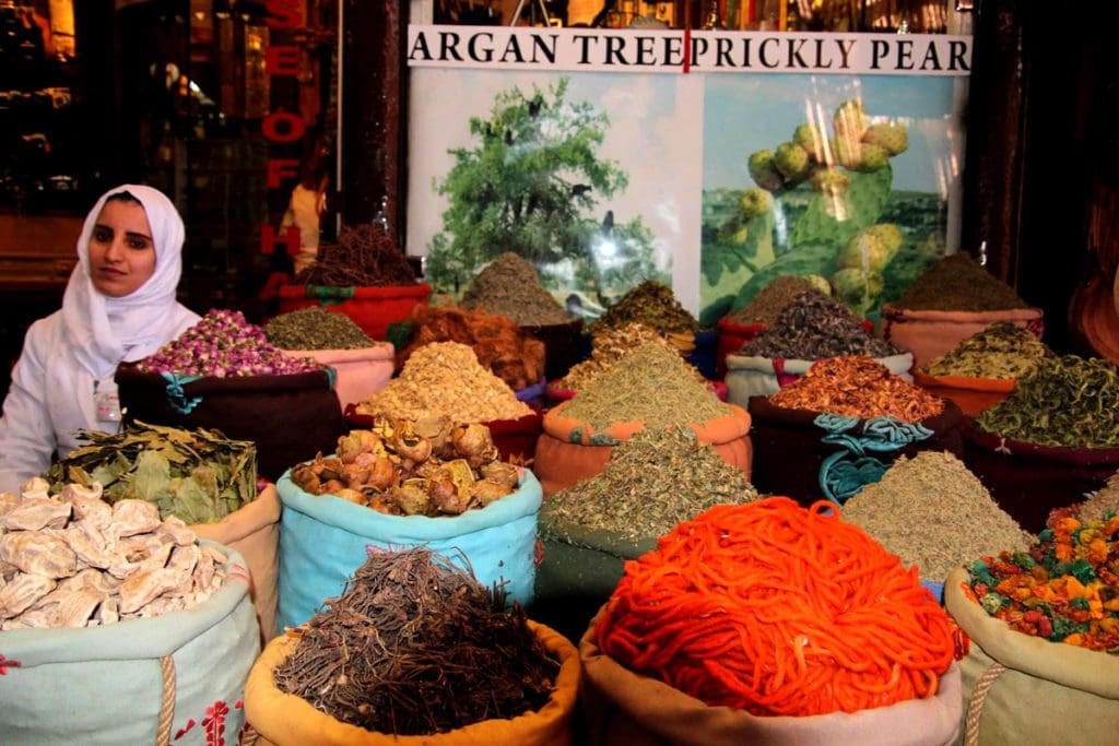 A woman sits selling spices at a spice market in Marrakesh, featuring several large heaps of colorful spices.