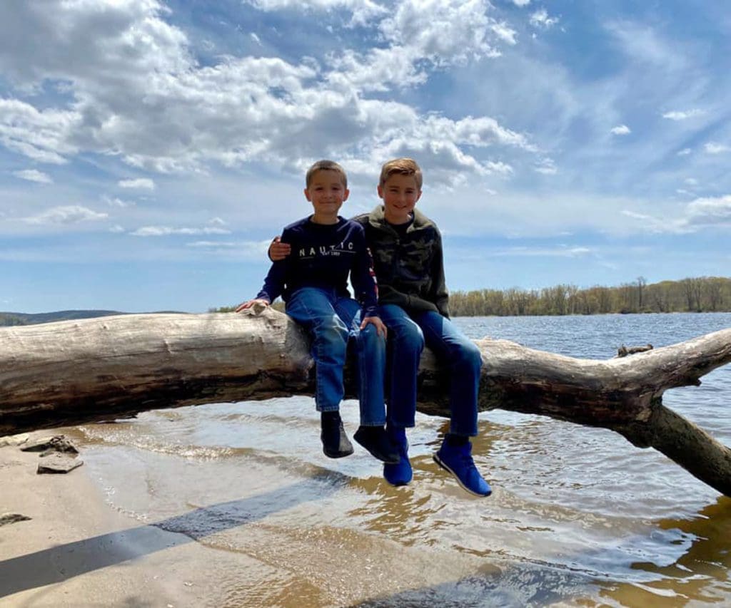 Two kids sit together on an overturned tree, reaching into the waters of a river at Gillette Castle State Park near Mystic, one of the best Labor Day Weekend getaways near NYC with kids.