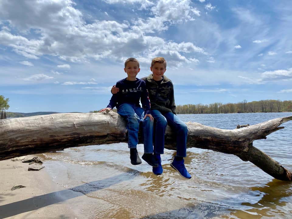 Two kids sit together on an overturned tree, reaching into the waters of a river at Gillette Castle State Park.