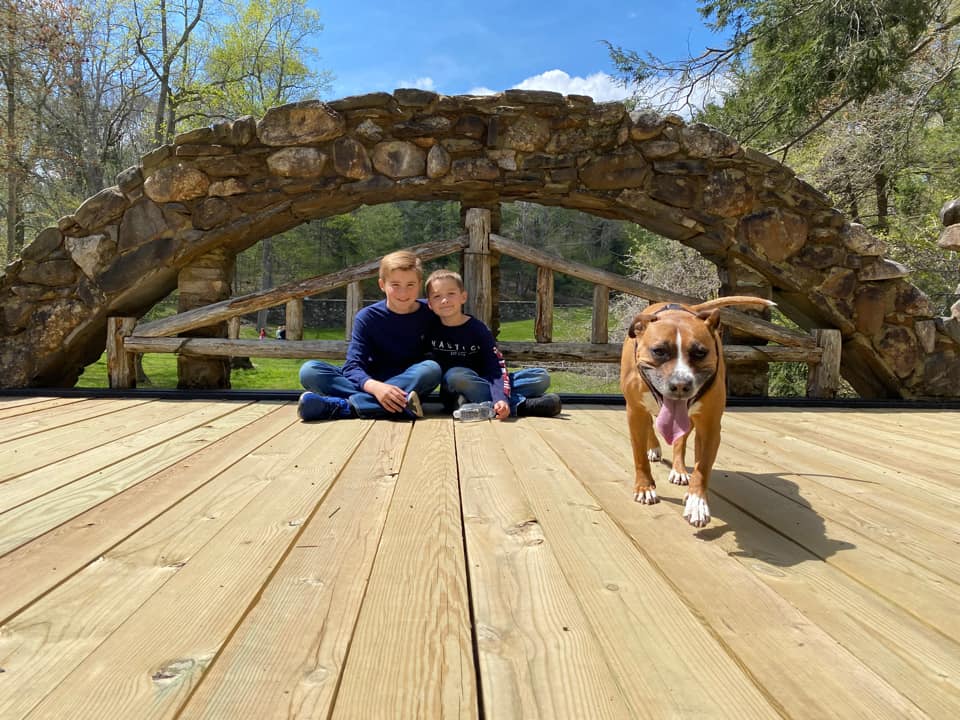 Two kids sit together on a platform, while a dog trots toward the camera, at Gilette Castle State Park.