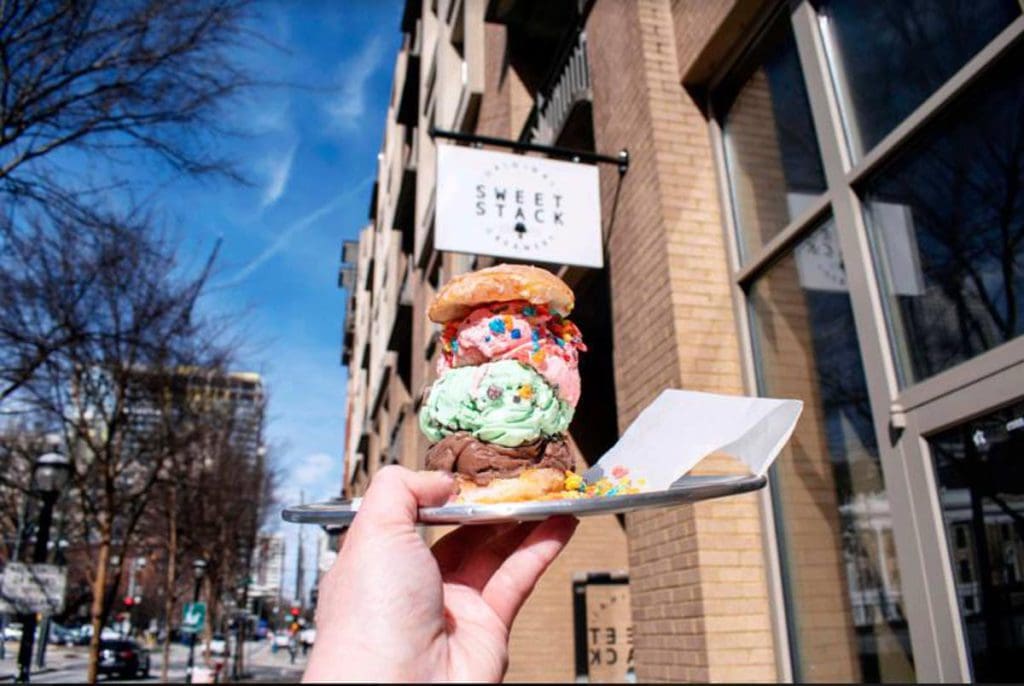 A hand holds out a stack of ice cream with the sign for Sweet-Stack Creamery in the distance.