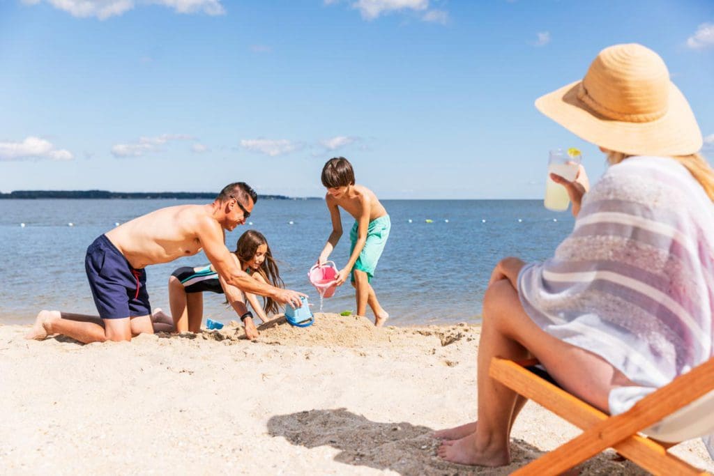 A mom watches from a beach lounger, while her husband helps two kids build a sand castle near the ocean in Williamsburg, one of the best places to visit during Memorial Day Weekend near NYC for families.