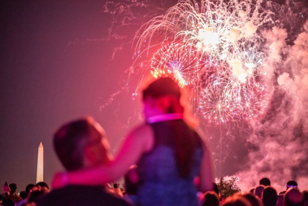 A man holds his young daughter while watching fireworks in DC, the Washington Monument is seen in the distance.