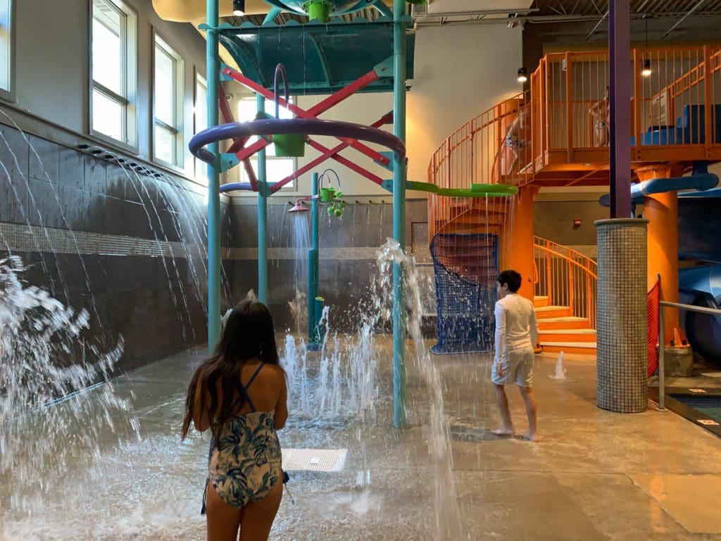 A young girl and boy, both wearing swim suits, run around the indoor splash pad area at Woodloch Resort.