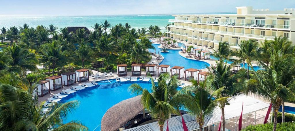 An aerial view of Azul Beach Resort Riviera Cancun, showcasing its large pool, white resort building, swaying palms around the grounds, and beachfront access.
