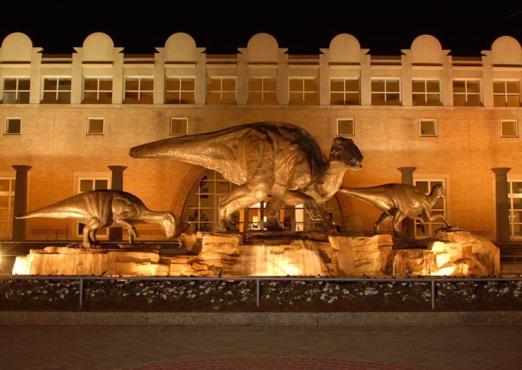 The exterior to the Fernbank Museum of Natural History at night, with its iconic triplet of dinosaur statues lit up.