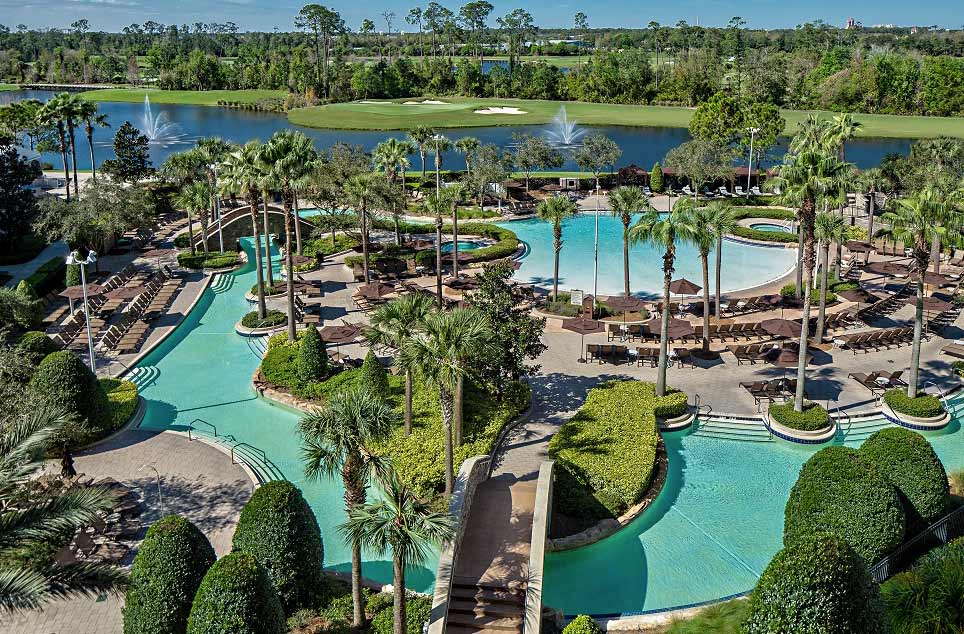 An aerial view of the outdoor swimming pools, and nearby pond, surrounded by swaying palms at Signia by Hilton Orlando Bonnet Creek.