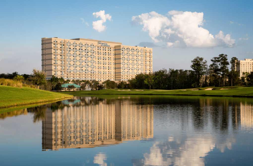 A large resort building of the Signia by Hilton Orlando Bonnet Creek stands proudly near a small body of water, reflecting in the pool.