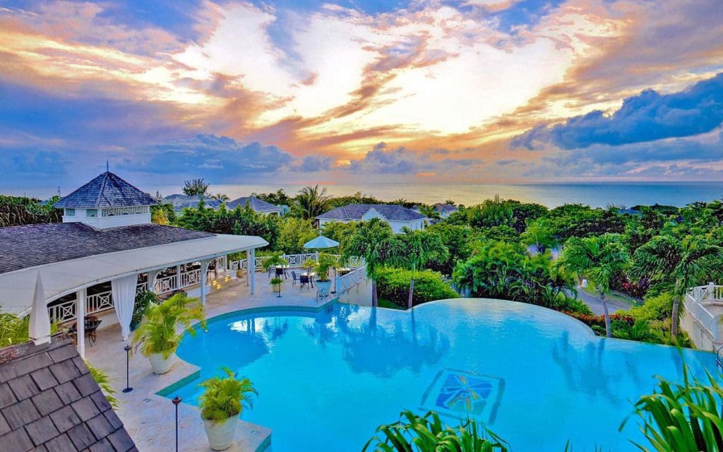 An aerial view of the pool, nearby pavilion, and lush grounds of Sugar Hill Estate, with the ocean in the distance.