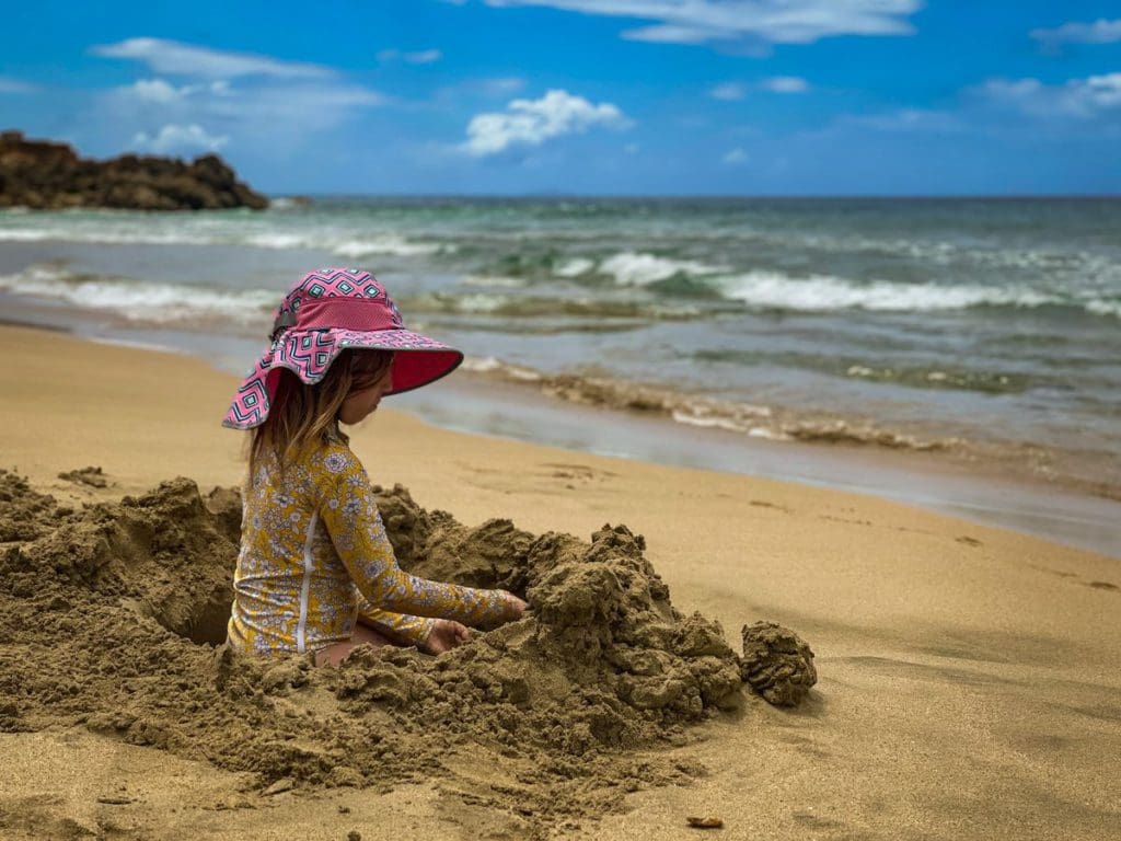 A young girl sits in a hole she dug in the stand, while wearing a wide-brimmed hat, a must on a beach packing list for families.