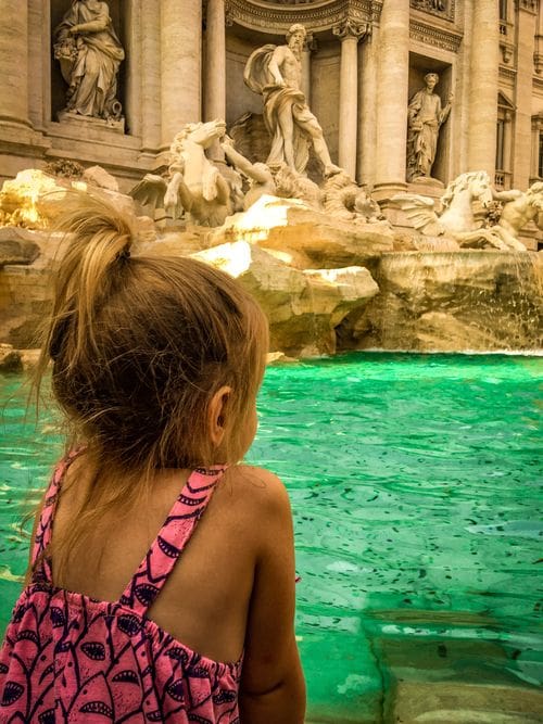 A young girl leans over the side of the Trevi Fountain to peer into the water, one of the best things to do Rome with toddlers.