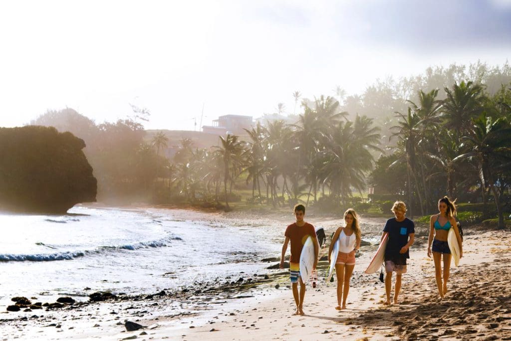 Four people, each holding surf boards, walk across a beach in Barbados. Barbados is a great place to go surfing with kids!