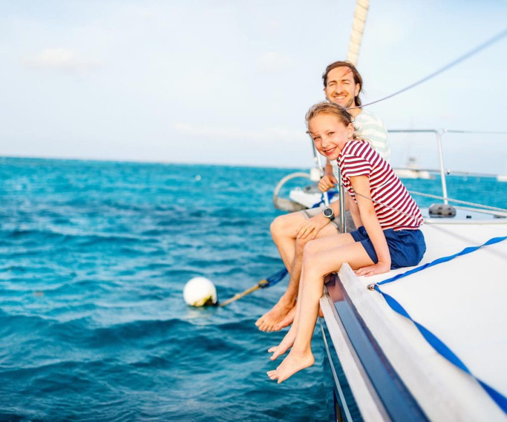 A dad and his daughter sit on the edge of a sail boat, with their legs hanging over the edge, above the water.