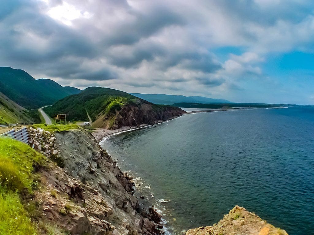 A striking shoreline featuring clear waters that meet rocky embankments at Cape Breton Island, with mountains in the distance.