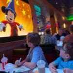 Two young boys look up at a screen, where Mickey Mouse is dancing, while eating dinner on a Disney Cruise Line.