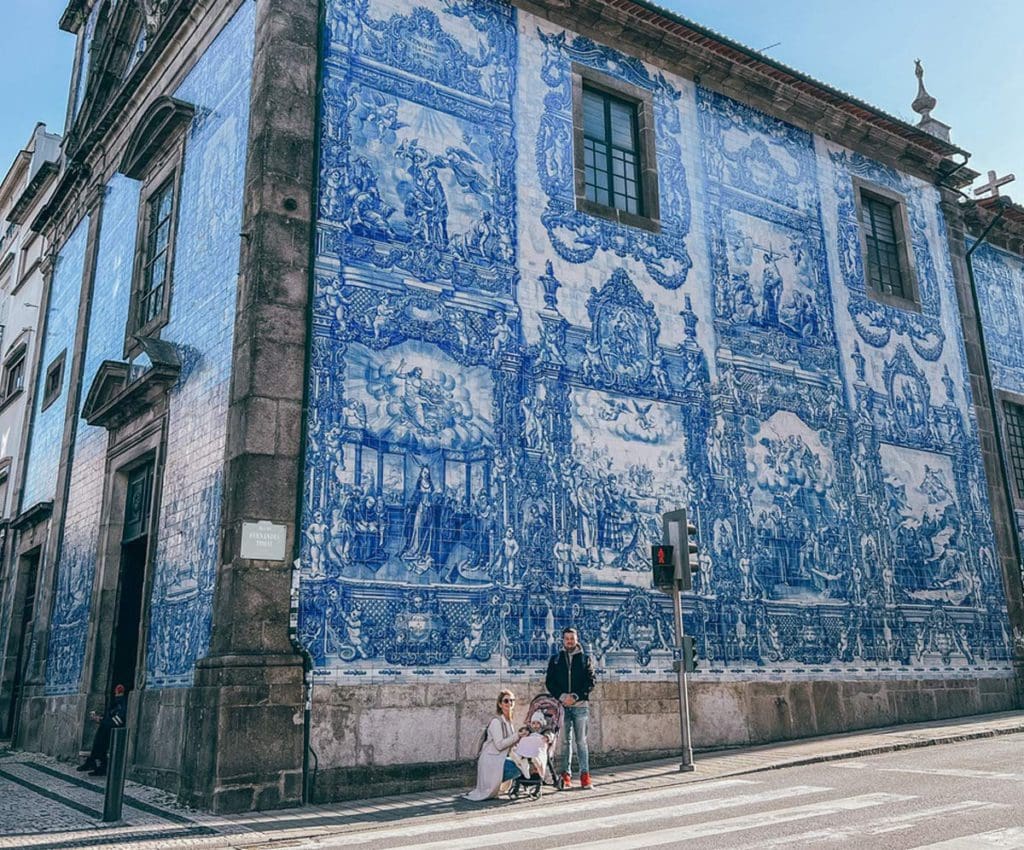 A family of three stands together in front of the Chapel of Souls, which features one of Porto's stunning building designs in blue and white.