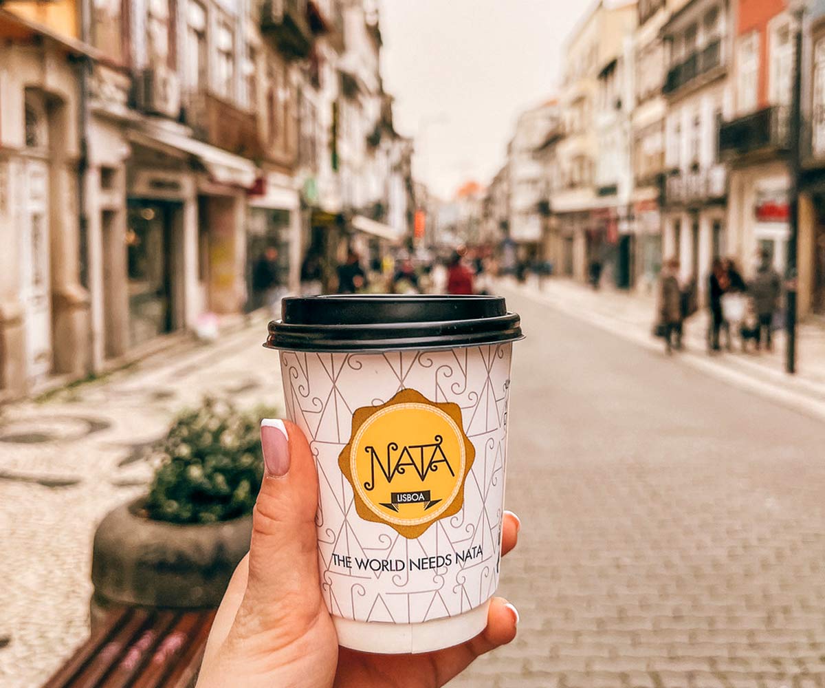 A hand holds a coffee from Nata with a Porto street in the distance.