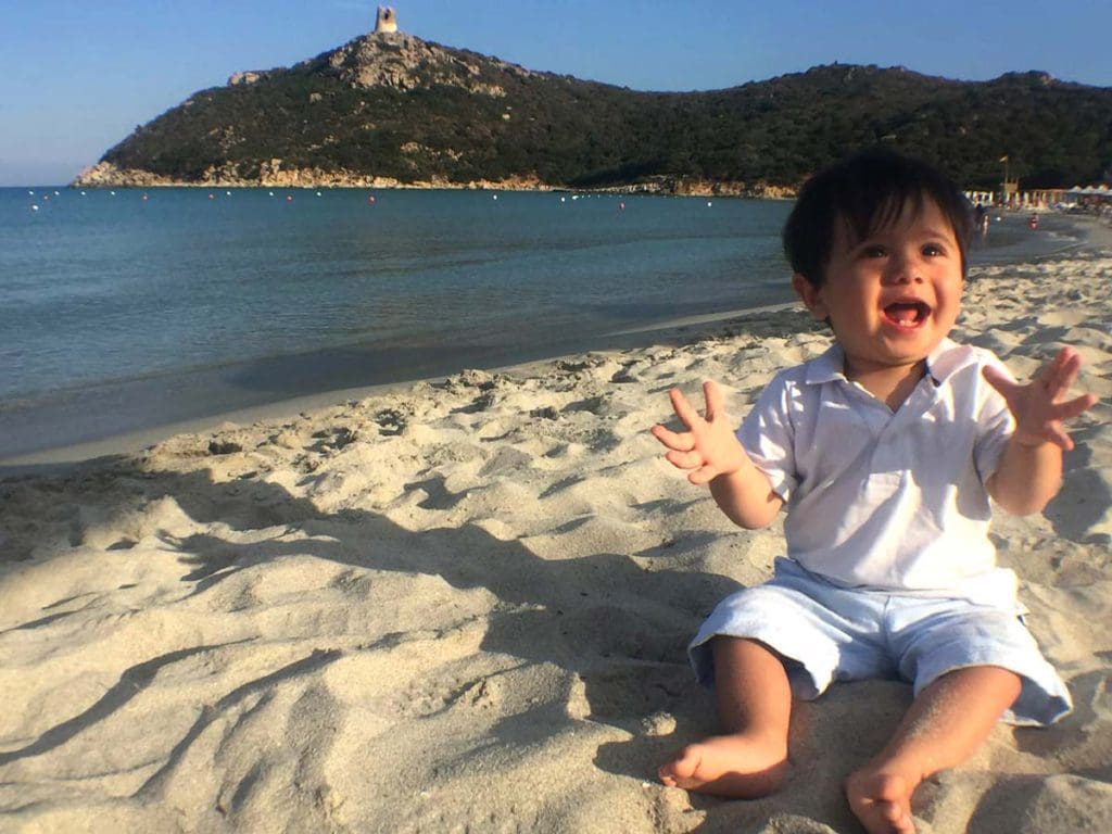 An infant boy claps his hands, while sitting on a beach in Sardinia, one of the best places to visit in Italy on a budget with kids. 