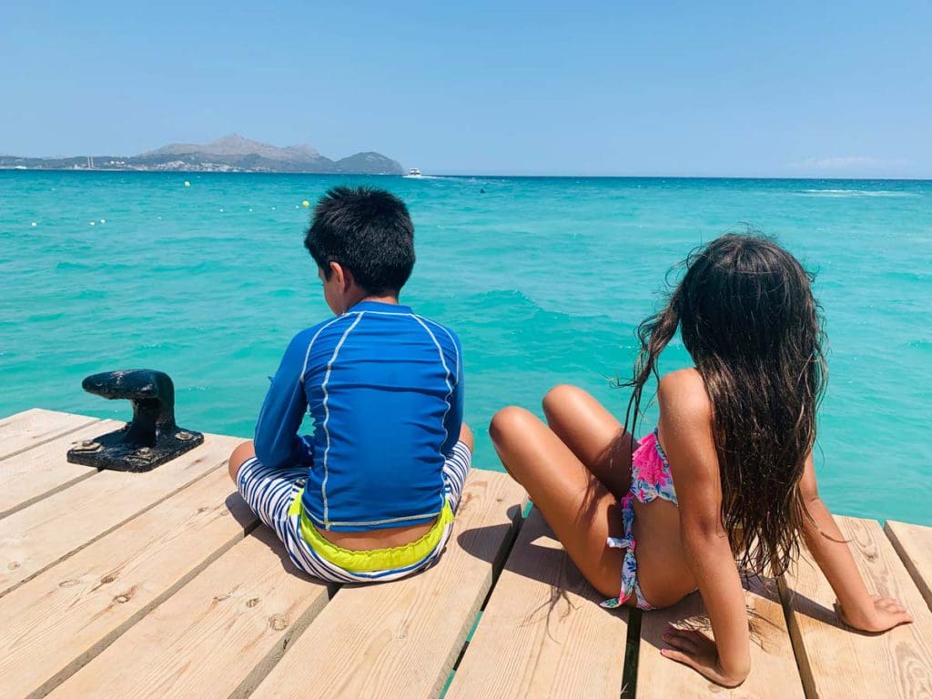 Two kids sit on a dock looking out onto a clear blue ocean in Mallorca.