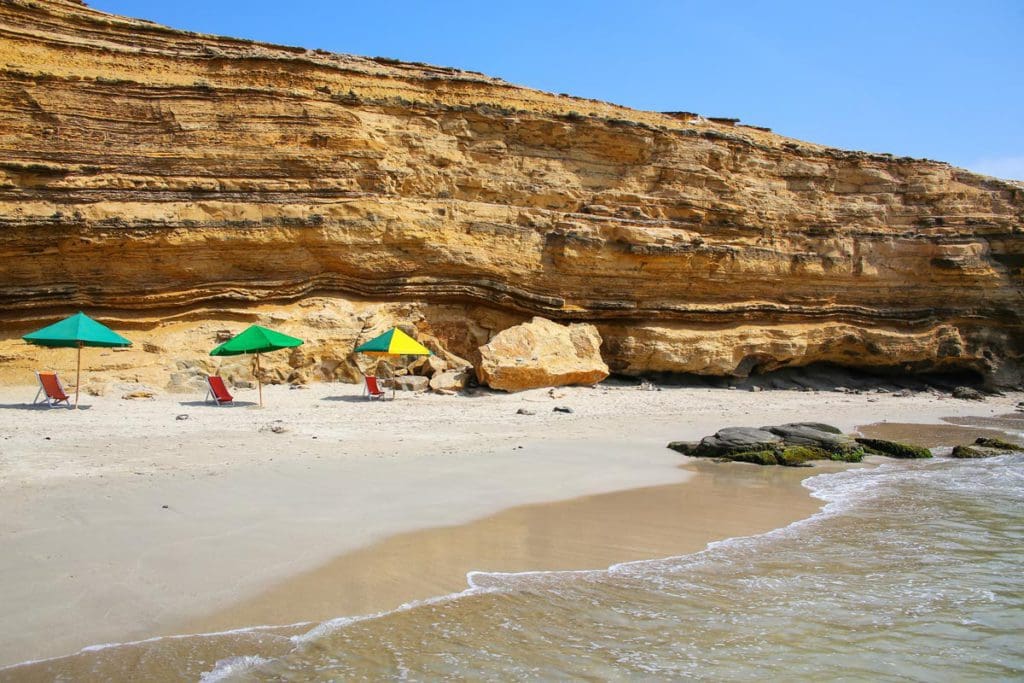 A beach, with umbrellas near the large stone wall in the distance, at Paracas, a great way to end our Peru family vacation itinerary.