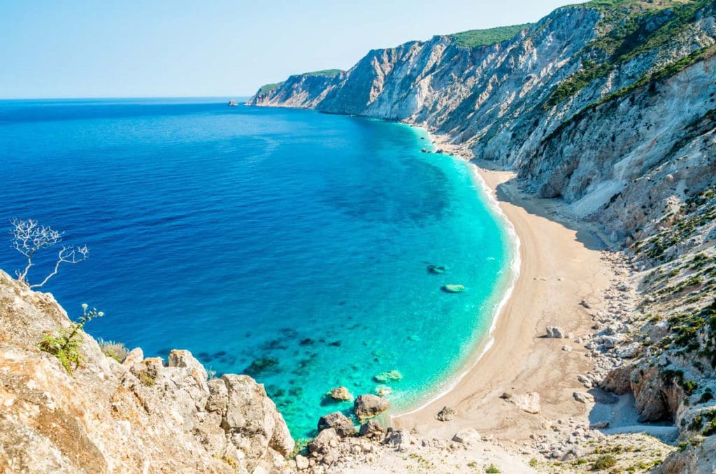 An aerial view of Platia Ammos Beach in Kefalonia, featuring turquoise waters and white sands, with a mountain raising from the sands.