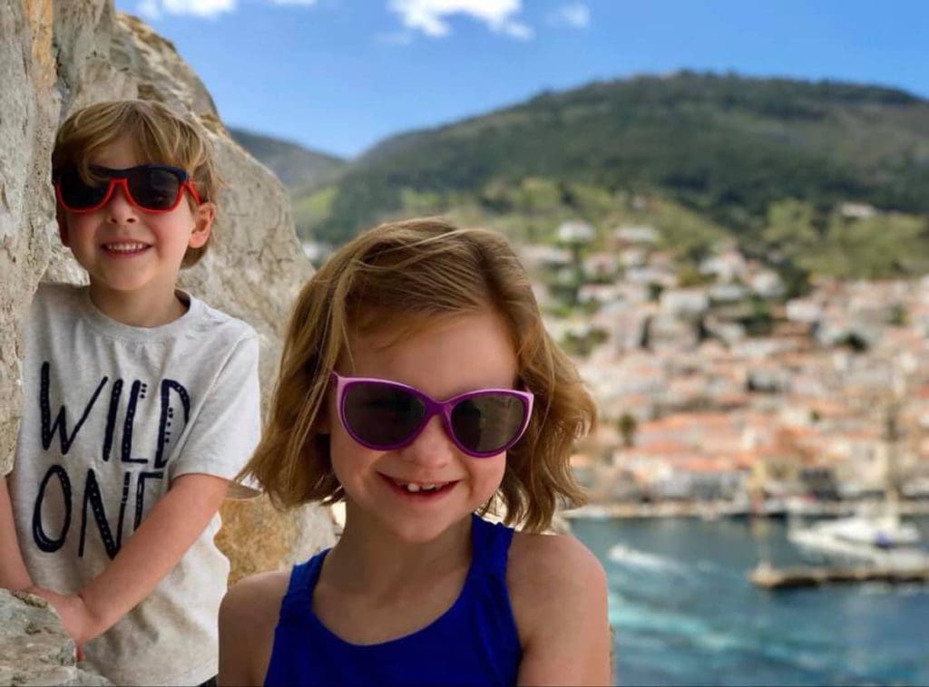 Two kids stand together and smile with a view of Idra, Greece in the distance, along the ocean.