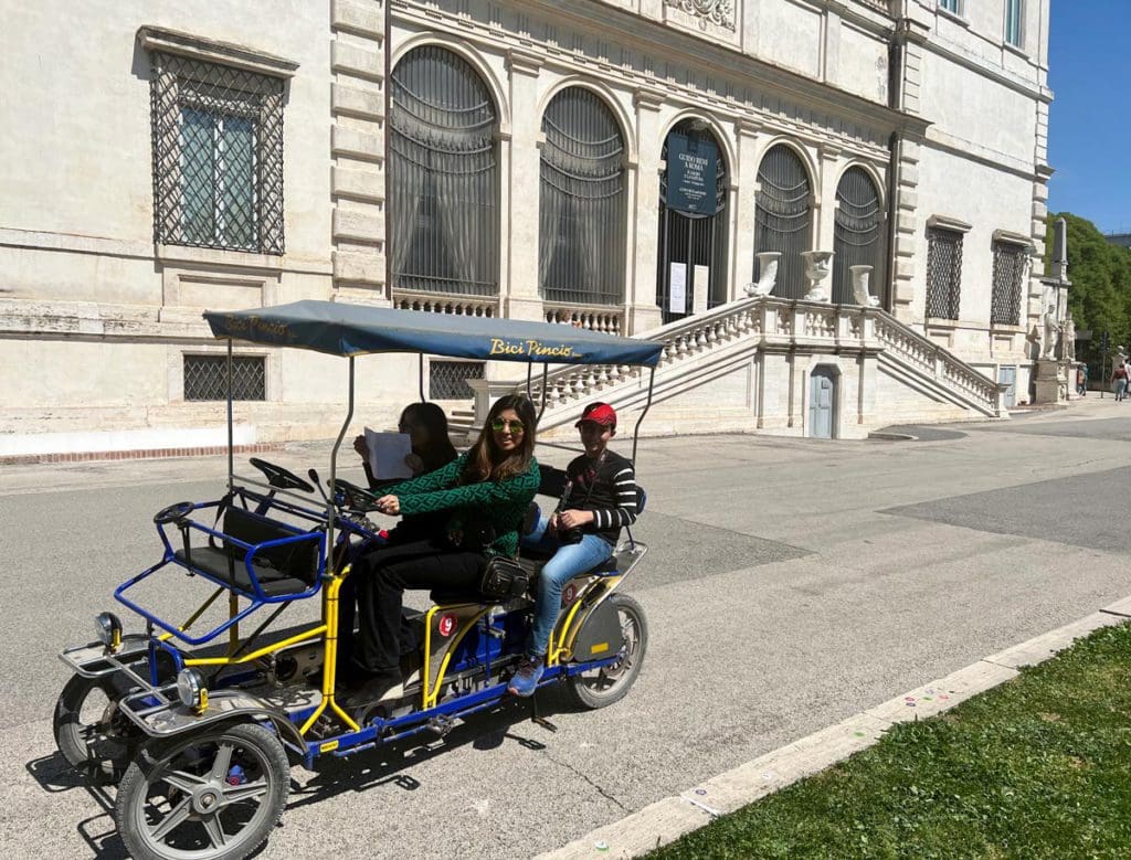 A family of four rides a surrey bike around Villa Borghese, with the museum behind them, this is one of the best neighborhoods to stay in Rome with kids.