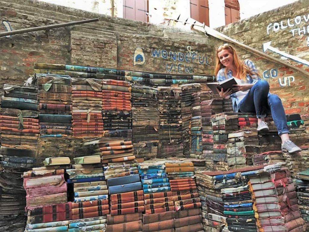 A woman leans against an outdoor display of books at the iconic Acqua Alta Bookshop, one of the best places to include on a Venice itinerary with kids. 