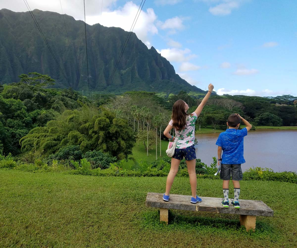 Two kids stand on a bench looking at the water while exploring Hoomaluhia Botanical Gardens.